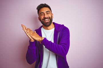 Young indian man wearing purple sweatshirt standing over isolated pink background clapping and applauding happy and joyful, smiling proud hands together