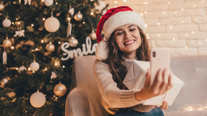 Woman in santa hat hugging presents and  taking selfie sitting on the floor near Christmas tree in the decorative interior. Christmas and New Year photo.