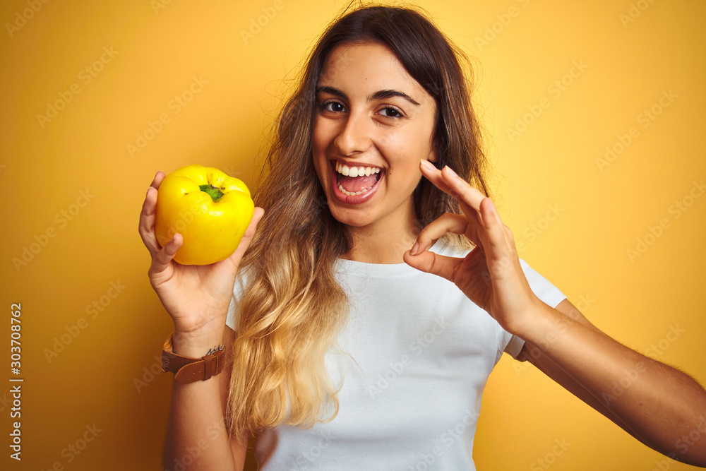 Sticker young beautiful woman holding pepper over yellow isolated background doing ok sign with fingers, exc