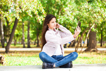 Brunette girl photographs herself on a cell phone while sitting in an autumn park.