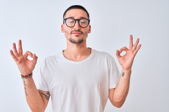 Young handsome man wearing glasses and standing over isolated background relax and smiling with eyes closed doing meditation gesture with fingers. Yoga concept.