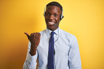 African american operator man working using headset over isolated yellow background smiling with happy face looking and pointing to the side with thumb up.