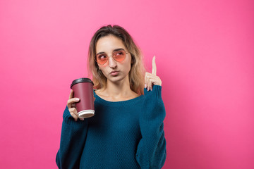 Glamor woman in glasses in an orange sweater with a drink of coffee on a pink background..	