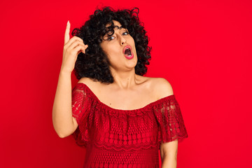 Young arab woman with curly hair wearing casual dress over isolated red background pointing finger up with successful idea. Exited and happy. Number one.