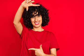 Young arab woman with curly hair wearing casual t-shirt over isolated red background smiling making frame with hands and fingers with happy face. Creativity and photography concept.