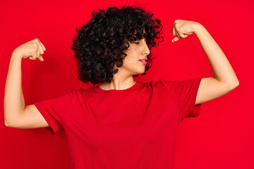 Fototapeta na wymiar Young arab woman with curly hair wearing casual t-shirt over isolated red background showing arms muscles smiling proud. Fitness concept.