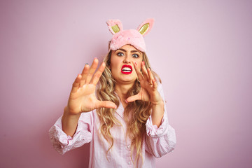 Young beautiful woman wearing pajama and sleep mask over pink isolated background afraid and terrified with fear expression stop gesture with hands, shouting in shock. Panic concept.
