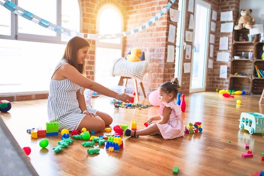 Young beautiful teacher and toddler sitting on the floor playing with building blocks toy at kindergarten
