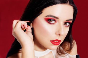Portrait, model holds mascara in hands and paints eyelashes. Beautiful brunette with professional red make-up, on a red background. New Year's red make-up