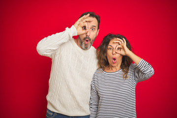 Beautiful middle age couple wearing winter sweater over isolated red background doing ok gesture shocked with surprised face, eye looking through fingers. Unbelieving expression.
