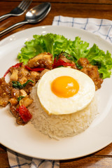 Fried basil chicken with fried egg and white rice in white dish on wooden table.