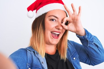 Beautiful woman wearing Christmas Santa hat make selfie over isolated white background with happy face smiling doing ok sign with hand on eye looking through fingers