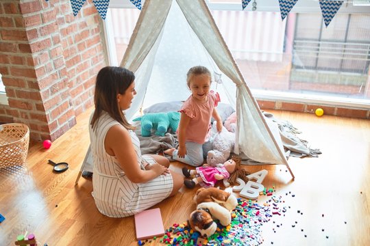 Beautiful teacher and blond toddler girl playing with dolls inside tipi at kindergarten