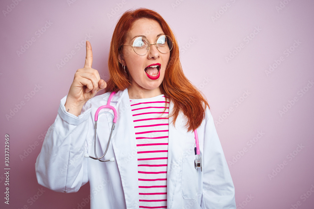 Sticker Young redhead doctor woman wearing glasses over pink isolated background pointing finger up with successful idea. Exited and happy. Number one.