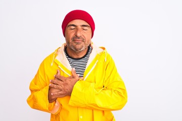 Middle age man wearing rain coat and woolen hat standing over isolated white background smiling with hands on chest with closed eyes and grateful gesture on face. Health concept.