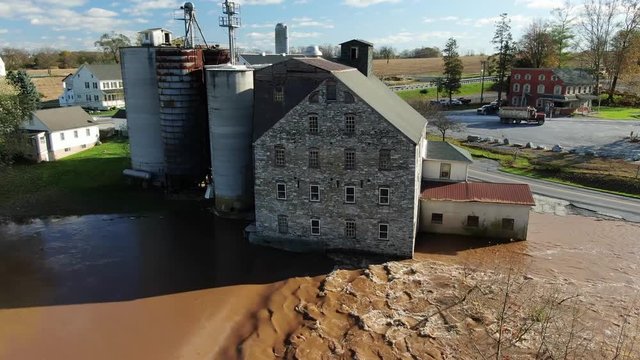 Aerial of flooded grain mill, silos, elevators beside muddy river on sunny day
