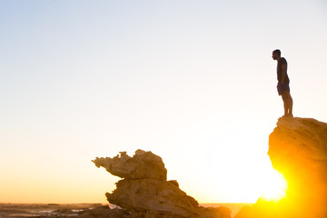 man standing on a rocky mountain cliff at golden hour wearing a black t-shirt and shorts with the golden sun rays peaking from underneath the rocks 