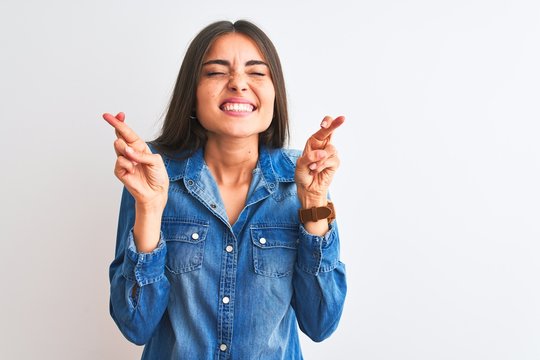 Young beautiful woman wearing casual denim shirt standing over isolated white background gesturing finger crossed smiling with hope and eyes closed. Luck and superstitious concept.