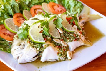Steam golden pompano fish with chili and lime - spicy thai food. Serve on white plate and decorate with lettuce, lime, garlic and tomato.
