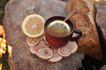 wooden cup with tea and lemon inside, with a wooden spoon, on a brown wooden plate, sliced ​​lemon next to it, on a stone