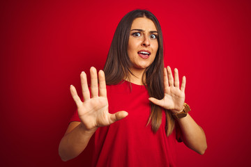 Young beautiful woman wearing t-shirt standing over isolated red background afraid and terrified with fear expression stop gesture with hands, shouting in shock. Panic concept.