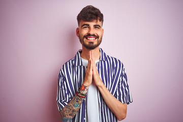 Young man with tattoo wearing striped shirt standing over isolated pink background praying with hands together asking for forgiveness smiling confident.