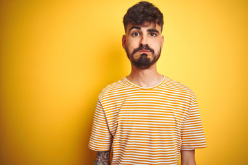 Young man with tattoo wearing striped t-shirt standing over isolated yellow background puffing cheeks with funny face. Mouth inflated with air, crazy expression.