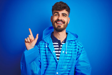 Young handsome man wearing rain coat standing over isolated blue background with a big smile on face, pointing with hand and finger to the side looking at the camera.