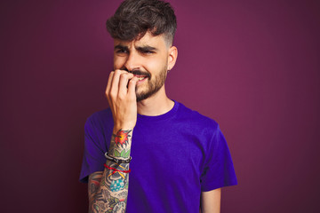Young man with tattoo wearing t-shirt standing over isolated purple background looking stressed and nervous with hands on mouth biting nails. Anxiety problem.