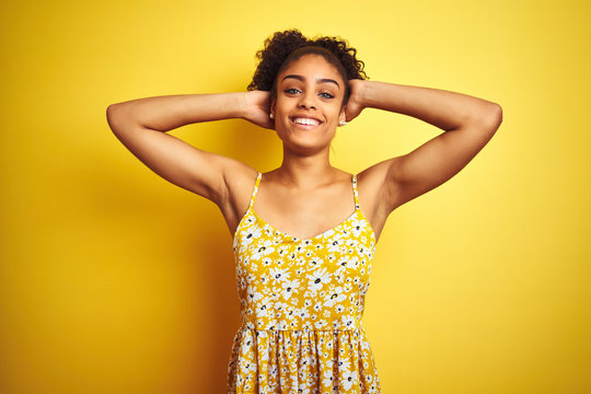 African american woman wearing casual floral dress standing over isolated yellow background relaxing and stretching, arms and hands behind head and neck smiling happy