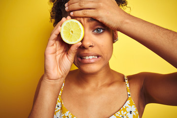 Young african american woman holding slice of lemon over isolated yellow background stressed with hand on head, shocked with shame and surprise face, angry and frustrated. Fear and upset for mistake.
