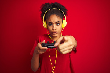 Afro woman playing video game using joystick and headphones over isolated red background pointing with finger to the camera and to you, hand sign, positive and confident gesture from the front