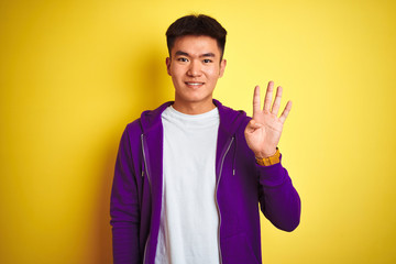Young asian chinese man wearing purple sweatshirt standing over isolated yellow background showing and pointing up with fingers number four while smiling confident and happy.