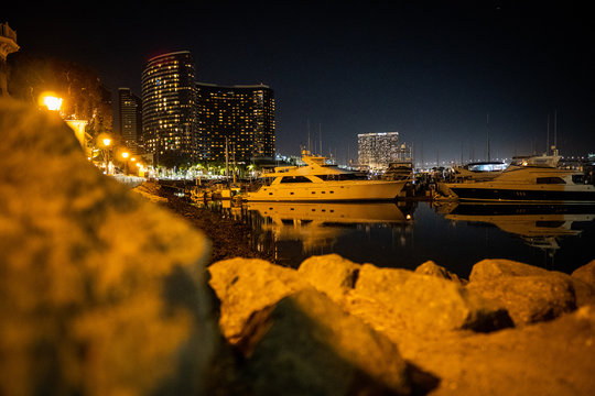 Image of Boats in San Diego California Marina with skyscrapers in the background at night with rocks in the foreground glowing from street lamps