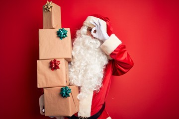 Senior man wearing Santa Claus costume holding tower of gifts over isolated red background with happy face smiling doing ok sign with hand on eye looking through fingers