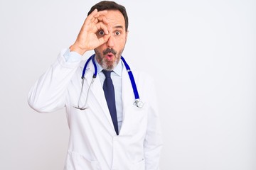 Middle age doctor man wearing coat and stethoscope standing over isolated white background doing ok gesture shocked with surprised face, eye looking through fingers. Unbelieving expression.