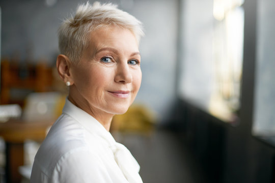 Close up portrait of attractive mature Caucasian woman with blonde pixie haircut and wrinkles looking at camera with confident smile while working in her office. Business, work, job and profession