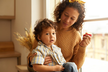 Indoor shot of joyful young female wearing sweater and headscarf eating apple on windowsill with adorable chubby baby boy on her lap. Family bonds, relationships, love and parenthood concept