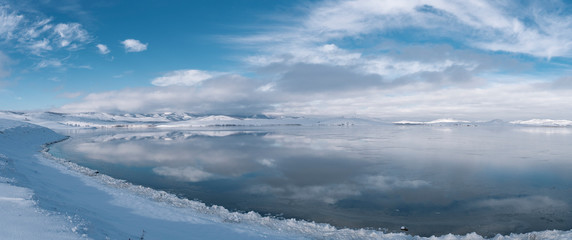 Panorama of winter landscape near Ural mountains; sunny snowy day with deep blue cloudy sky on freezing lake with hills reflection on water surface; season changing in wild nature, Russia