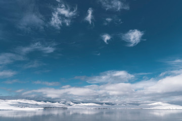 Panorama of winter landscape near Ural mountains; sunny snowy day with deep blue cloudy sky on freezing lake with hills reflection on water surface; season changing in wild nature, Russia