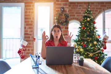 Beautiful woman sitting at the table working with laptop at home around christmas tree looking surprised and shocked doing ok approval symbol with fingers. Crazy expression