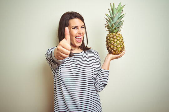 Young beautiful woman holding tropical fruit pineapple over isolated background happy with big smile doing ok sign, thumb up with fingers, excellent sign
