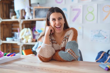 Young beautiful teacher woman wearing sweater and glasses sitting on desk at kindergarten with a...