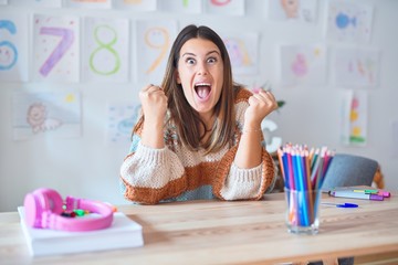 Young beautiful teacher woman wearing sweater and glasses sitting on desk at kindergarten...