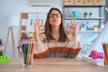 Young beautiful teacher woman wearing sweater and glasses sitting on desk at kindergarten relaxed and smiling with eyes closed doing meditation gesture with fingers. Yoga concept.