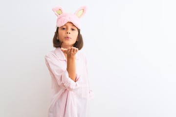 Beautiful child girl wearing sleep mask and pajama standing over isolated white background looking at the camera blowing a kiss with hand on air being lovely and sexy. Love expression.