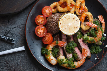Close-up of surf and turf with marbled beef meat, shrimps, calamari rings, tomatoes and chumichurri, above view