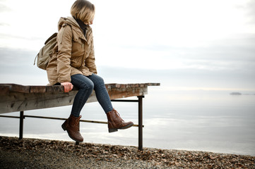 young girl in a warm jacket sits on the pier with his legs dangling and look towards the sea
