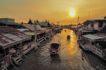 Obraz premium view evening of long-tail boats running in Amphawa Canal around with wooden houses and yellow sun light in the sky background, sunset at Amphawa Floating Market, Samut Songkhram, Thailand.