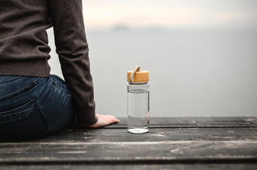 glass reusable bottle for water, stands on a wooden pier against the background of a cloudy sky next to a sitting figure of a girl